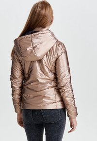 Vrouw met lang haar, gekleed in een glanzende lichtbruine gewatteerde jas met capuchon en donkere spijkerbroek, staande met haar rug naar de camera tegen een effen achtergrond.