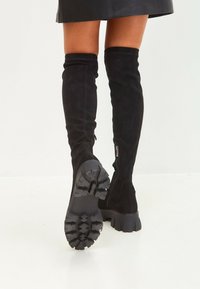 Zwarte boven-de-knie laarzen van suède met een gestructureerde rubberen zool en een zijritsdetail. De hak is chunky en robuust.