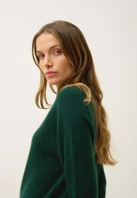 Ciemnozielony sweter z miękkiego, teksturowanego materiału o luźnym kroju. Widać długie rękawy i okrągły dekolt; tło jest gładkie i neutralne.