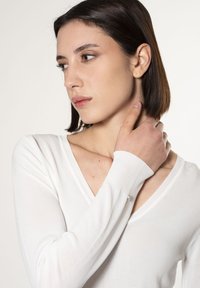 Weißer, langärmliger Pullover mit tiefem V-Ausschnitt, gerippten Bündchen und zarten Akzentdetails nahe dem Handgelenk. Weicher, leichter Stoff.