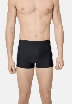 Torso y piernas masculinos con bañador negro con franjas blancas laterales, de pie frente a un fondo claro y liso.