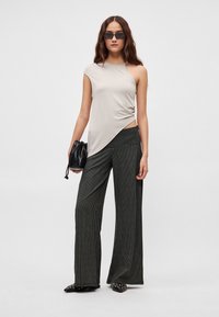 Asymetryczna beżowa bluzka z miękkiego materiału w połączeniu z czarnymi, szerokimi spodniami w pionowe paski; modelka trzyma małą czarną torebkę.