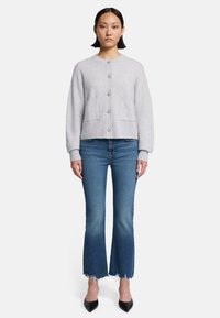 Cardigan grigio chiaro abbottonato con orlo a coste e due tasche frontali, indossato sopra jeans a zampa d'elefante blu e décolleté nere a punta.
