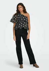 Femme aux longs cheveux bouclés portant un top noir asymétrique à pois blancs, un jean noir et des bottines marron.