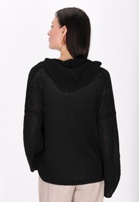 Pulover cu glugă tricotat negru, cu o croială lejeră, având un material texturat și mâneci late. Spatele este neted, cu un decolteu relaxat.