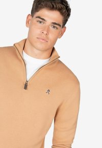Sudadera cremosa con cierre y cuello alto, que presenta un logo sutil en azul marino. La tela parece lisa y tiene una textura suave.