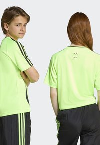 Neon žalia sportinė marškinė su juodais akcentais, trumpomis rankovėmis, apvaliu kaklu ir logotipu ant nugaros. Dėvimi su juodomis kelnėmis su juostelėmis.