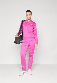 Pink tracksuit made of smooth, lightweight fabric, featuring white stripes on the sleeves and pants. Paired with a black sports bag.