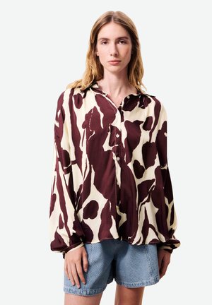 Femme portant une chemise ample à manches longues boutonnée avec un imprimé abstrait marron et crème, ainsi qu'un short en denim bleu clair, debout devant un fond uni.