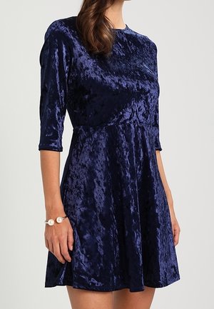 Kobieta ubrana w granatową sukienkę z karbowanego weluru do kolan, z rękawami do łokcia i perłową bransoletką na lewej ręce.