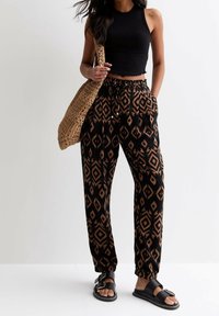 Femme portant un crop top sans manches noir, un pantalon à motifs noirs et bruns, des sandales noires, tenant un sac à épaule en toile tan tissée.