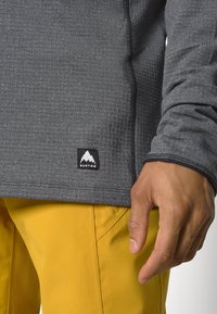 Camicia grigia a maniche lunghe con tessuto testurizzato, caratterizzata da un'etichetta nera con un logo di montagna bianco. Pantaloni gialli visibili nella parte inferiore.