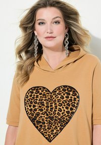 Haut à capuche beige avec un grand motif de cœur en imprimé léopard, entouré de noir. Texture lisse avec des manches courtes.