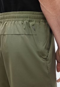 Gros plan sur une personne portant un pantalon vert olive avec une ceinture élastique et une poche latérale zippée, son bras droit visible à côté de la hanche.