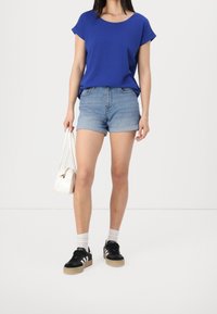 Nainen, jolla on yllään sininen lyhythihainen paita, vaaleat farkkushortsit, mustat tennarit, valkoiset sukat ja joka pitää valkoista käsilaukkua sivullaan.