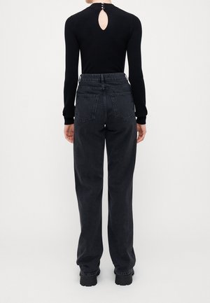 Person trägt ein schwarzes, geripptes Langarmoberteil mit Ausschnitt und Knopfdetail am Hals, kombiniert mit hoch taillierten, lockeren schwarzen Jeans und Stiefeln.