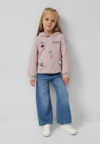 Dziewczyna stoi przy ścianie, ubrana w różową bluzę z kapturem z wzorem baletnic i napisem, niebieskie szerokie dżinsy oraz białe trampki.