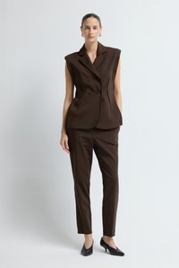 Vrouw in een mouwloze, donkerbruin, dubbele-breasted blazer met bijpassende broek en zwarte kitten heels, staand voor een effen witte achtergrond.