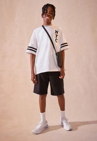 Baltas per dydį didelis marškinėlis su užrašu „NYC“, juodi šortai ir balti sportbačiai. Marškinėliai turi juodas juostas ant rankovių ir laisvalaikio dizainą.