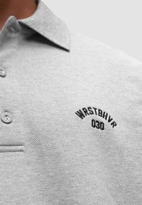 Szary polo z teksturowanej tkaniny, z kołnierzykiem, trzema guzikami i wyhaftowanym czarnym logo "WRSTBHV 030" na klatce piersiowej.