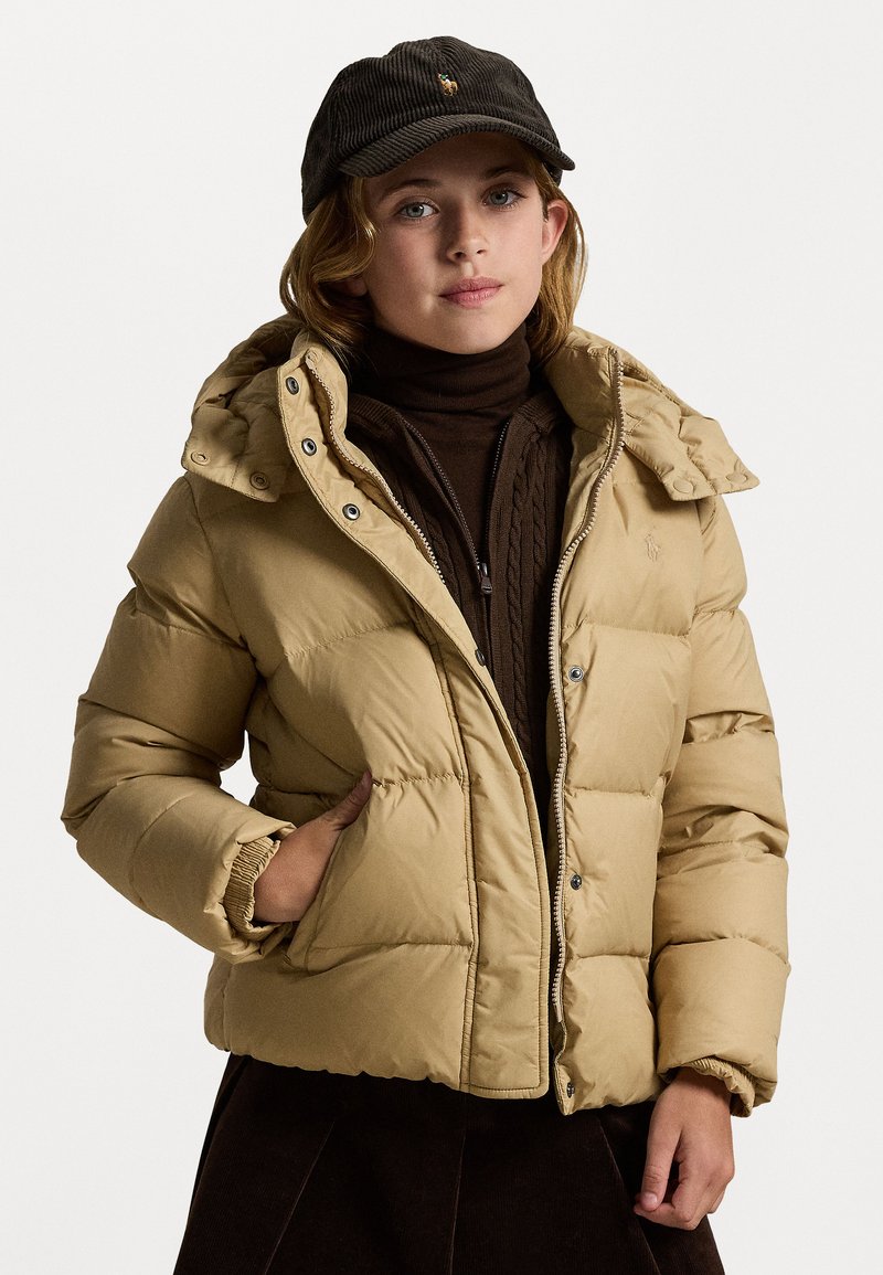 Puffjakke i lys tan med hette, med glidelås foran, sidelommer og ribbestrikkede mansjetter. Bært over en mørk brun turtleneck.