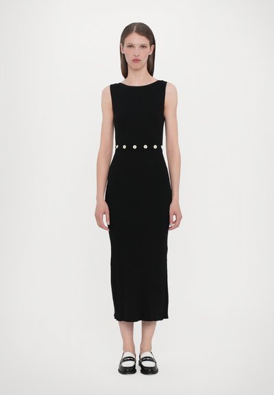 Schwarzes geripptes Strickkleid mit einer figurbetonten Silhouette und knielangem Schnitt, akzentuiert durch einen Gürtel mit großen weißen Knöpfen. Flache schwarze Schuhe.