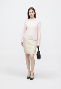 Hellrosa gestrickter Pullover mit langen Ärmeln, kombiniert mit einem engen cremefarbenen Rock aus Kunstleder. Schwarze Handtasche und Absatzschuhe runden den Look ab.