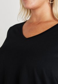 Črna majica s kratkimi rokavi v ovratniku v obliki črke V, narejena iz gladkega materiala. Ohlapen kroj, preprost dizajn in minimalno okrasje.