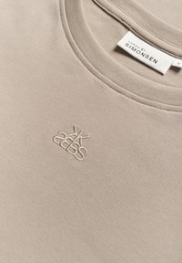Beige puuvillane sviiter, millel on ümmargune kaelus, tekstuuriga soonikkrae ja väike tikkitud logo rinnal.