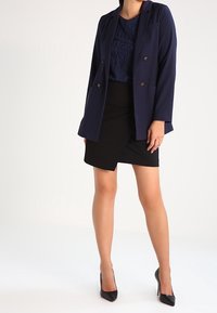 Blazer de doble botonadura en navy, blusa texturizada azul, falda envolvente negra y tacones negros puntiagudos. Apto para entornos profesionales.