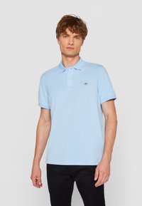 Šviesiai mėlynas polo marškinėlis, pagamintas iš medvilnės mišinio, su trumpomis rankovėmis, klasikine apykakle ir subtiliu logotipo akcentu krūtinėje.