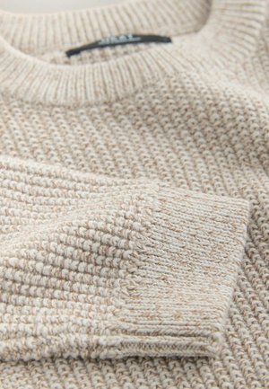 Neulottu neule kevyeen beigeen sävyyn, jossa on teksturoitu kuvio. Siinä on ribbineulottu pyöreä pääntie ja hihansuut, jotka korostavat pehmeää ja mukavaa kangasta.