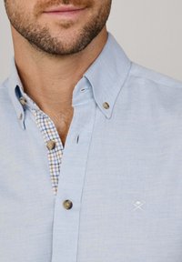 Camisa de botón azul claro con una tela texturizada, cuello interior a rayas en contraste y logo bordado en el pecho.