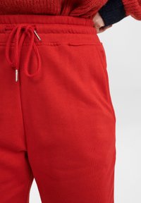 Närbild av röda svettbyxor med dragsko i midjan och sidofickor, bärs med en röd och svart tröja.