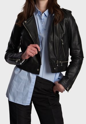 Personne portant une veste en cuir noire par-dessus une chemise bleu clair boutonnée et un pantalon noir, une main ajustant le zip de la veste.