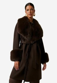 Cappotto di lana marrone con colletto in pelliccia, dettagli sfrangiati e polsini in pelliccia. Cintura in vita annodata che valorizza il design. Tessuto liscio e forma strutturata.