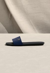 Σαγιονάρα σε ναυτικό μπλε με φαρδιά υφασμάτινη λωρίδα και μικρό χρυσό διακοσμητικό λογότυπο πάνω σε ανοιχτή ουδέτερη επιφάνεια.