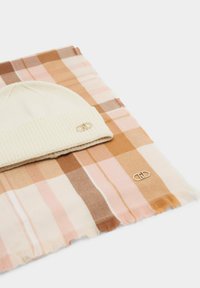 Cuffia in maglia color crema con polsino a coste e logo, posta sopra a una sciarpa a quadri nei toni del rosa tenue, marrone e beige con bordi sfrangiati.