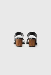 Chaussures à dos ouvert en cuir noir avec une large sangle et un talon en bois carré. Le design présente une texture lisse et une forme incurvée.