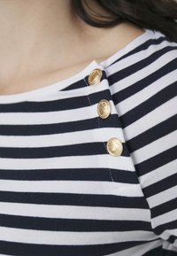 Marinblå och vit randig skjorta med rundad halsringning, som har tre gyllene dekorativa knappar vid axeln. Mjuk, elastisk tyg.