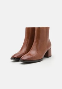 Bottes ankle en cuir marron avec une finition brillante, des pointes effilées et un talon épais. Présente un minimum de coutures et une fermeture éclair sur le côté.