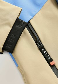 Beige og blå jakke med en glat tekstur og en sort lynlås med en label, der siger "BAKER." Synlig syning og detaljer.