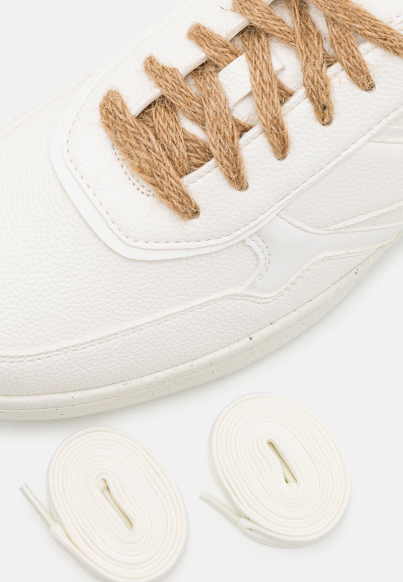 Witte leren sneaker met een gestructureerd oppervlak en bruine jute veters. Onder de sneaker liggen nog twee extra witte veters, één plat en één rond.