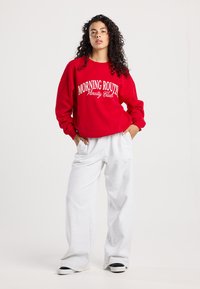 Rode sweatshirt met de opdruk "MORNING ROUTINE Varsity Club", gecombineerd met grijze joggingsbroek. Beide kledingstukken hebben een relaxed fit en een casual stijl.