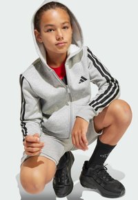 Grå zip-hoodie med svarta accenter, tajta shorts och svarta sneakers. Har tre svarta ränder på ärmarna. Modellen poserar på knä.