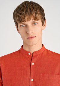 Camicia rossa con colletto, realizzata in tessuto strutturato; presenta una piccola patta con bottoni e una tasca sul petto. Nessun motivo o grafica visibile.