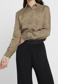 Olivově zelená saténová košile s špičatým límečkem, zapínáním na knoflíky a dlouhými rukávy, spárovaná s černými kalhotami s vysokým pasem. Měkká textura.