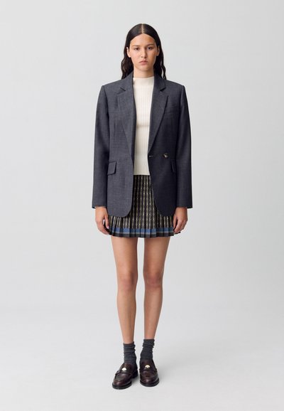 Blazer gris avec des revers crantés, pull blanc à côtes, et jupe plissée avec un motif de chaîne dorée, accompagnés de mocassins foncés et de chaussettes chevilles.
