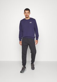 Felpa navy con scollo rotondo e piccolo logo bianco, abbinata a pantaloni jogger neri e scarpe da ginnastica grigie, in piedi su uno sfondo bianco.