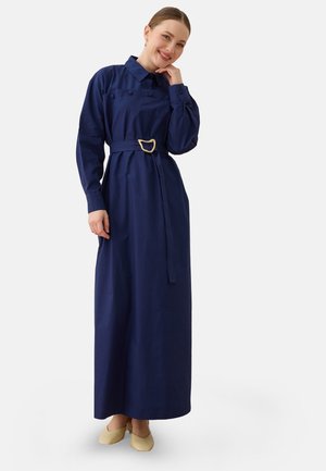 Donna sorridente che indossa un lungo vestito blu navy con cintura e maniche lunghe e scarpe beige basse, in piedi davanti a uno sfondo semplice.